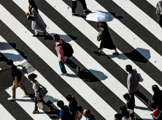 A flock of pedestrians crosses a large sunlit crosswalk